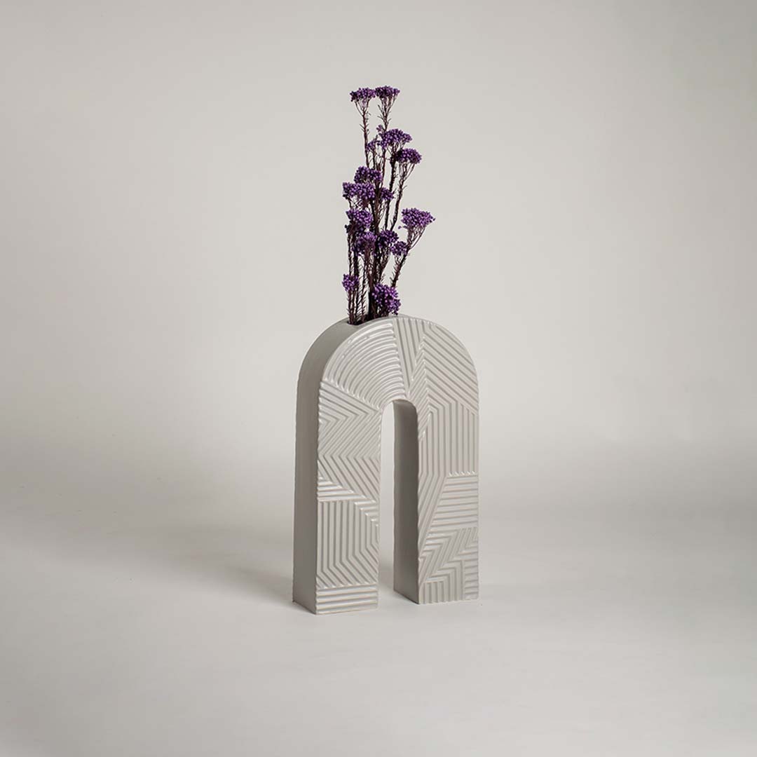 White ceramic arch vase with geometric texture holding purple flowers on a wood table next to a boucle chair for modern decor.