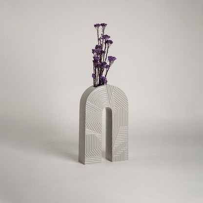 White ceramic arch vase with geometric texture holding purple flowers on a wood table next to a boucle chair for modern decor.