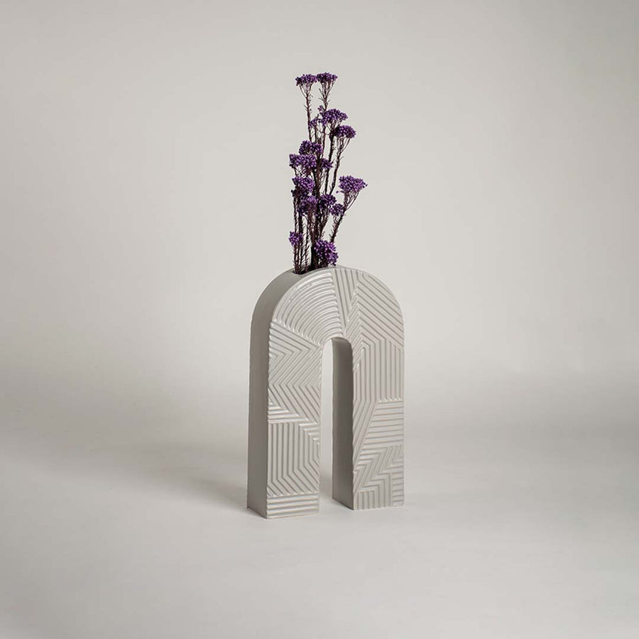 White ceramic arch vase with geometric texture holding purple flowers on a wood table next to a boucle chair for modern decor.