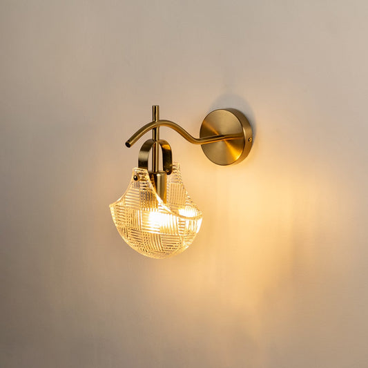 A gold metal wall light with a patterned clear glass shade shown with lights on against a white background highlighting its warm soft glow and elegant shape.