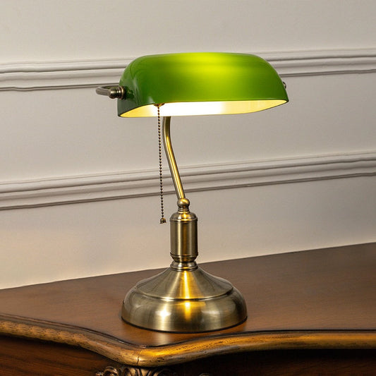 Green glass banker table lamp with antique brass base glowing softly on white background for vintage office decor.