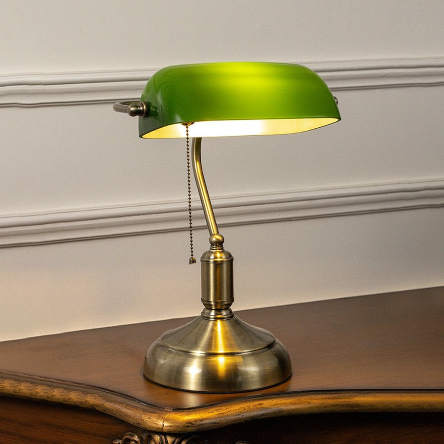 Green glass banker table lamp with antique brass base glowing softly on white background for vintage office decor.