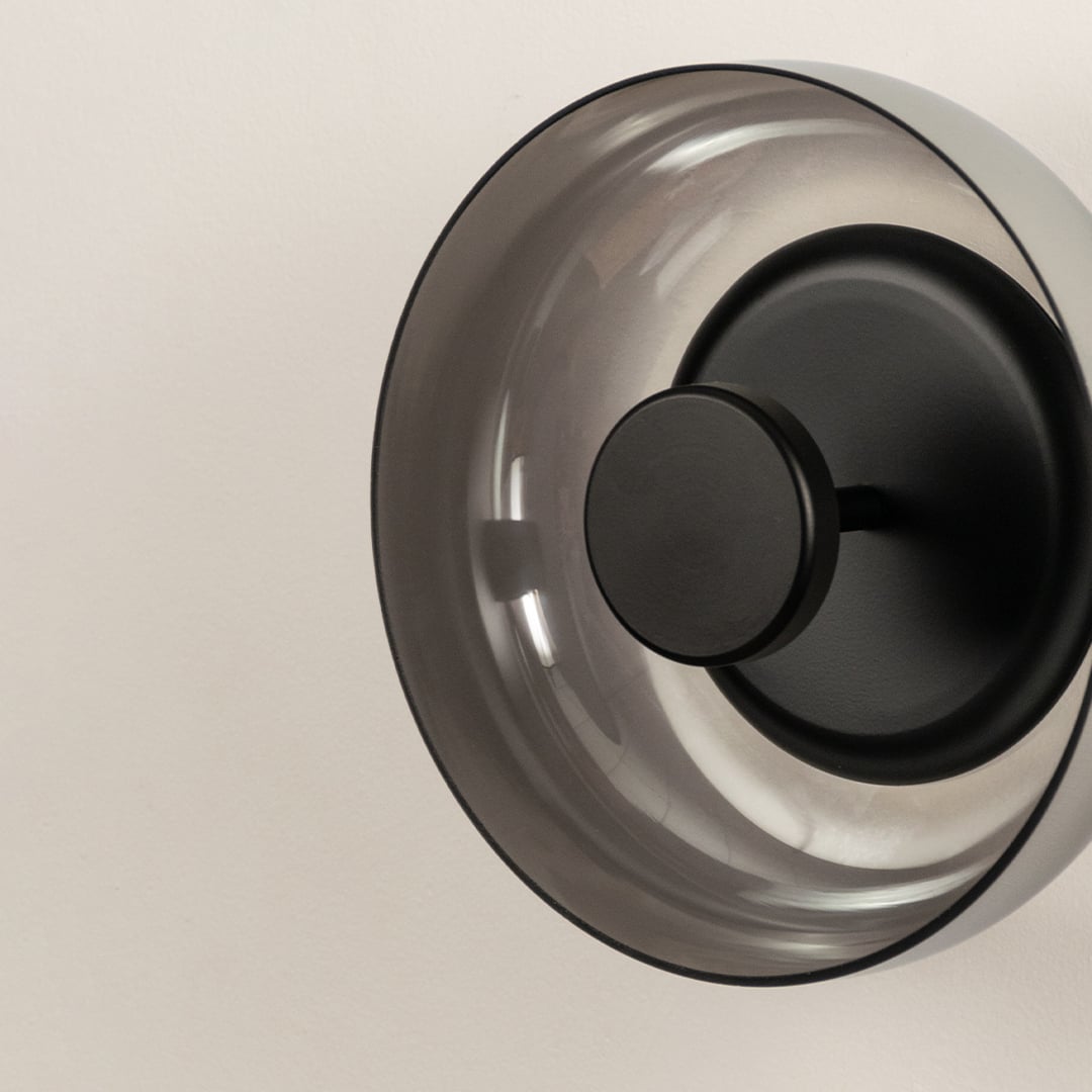 Detailed macro view of a contemporary round smoke glass wall lamp showcasing the matte black metal centerpiece mount.