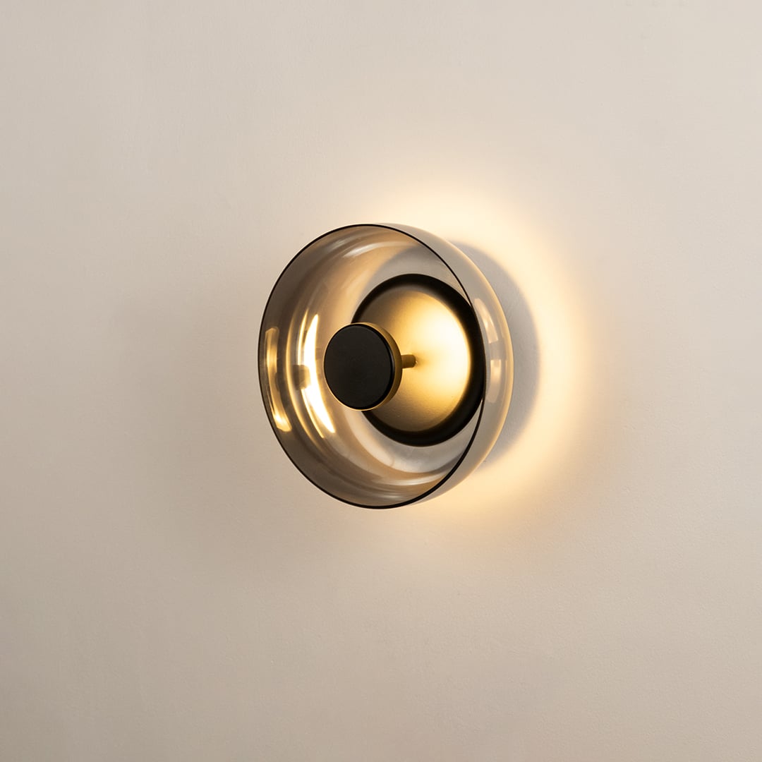 Modern circular smoke glass wall sconce turned on against a plain white background to display a warm halo backlight.