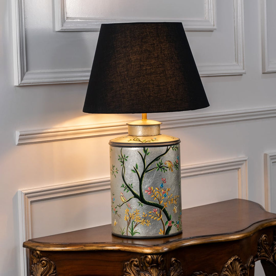 Table lamp on white background displaying ceramic base with hand painted botanical design accented by a classic black lampshade.