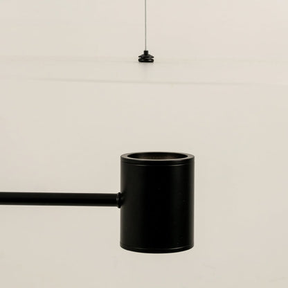 Close up of a black cylindrical ceiling light fitting with a slim rod attached shown against a light background with a thin hanging wire above.