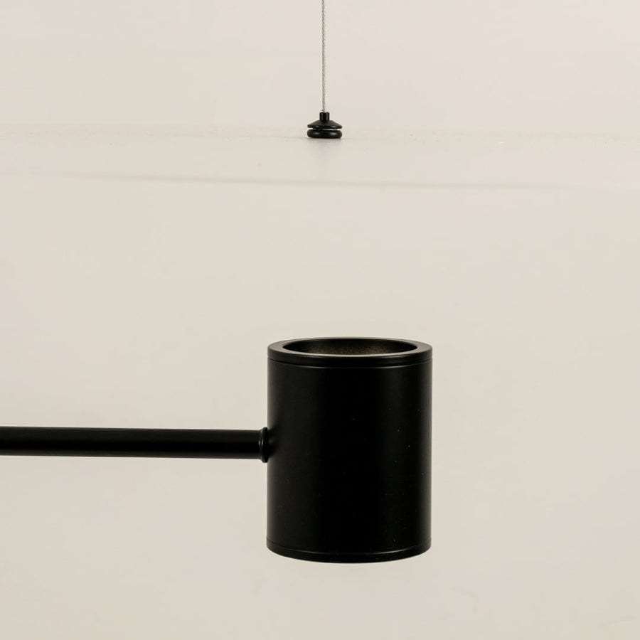 Close up of a black cylindrical ceiling light fitting with a slim rod attached shown against a light background with a thin hanging wire above.