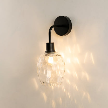 A black metal and clear glass wall light glowing warmly against a smooth pale wall spreading patterned reflections and adding stylish contemporary interior décor.