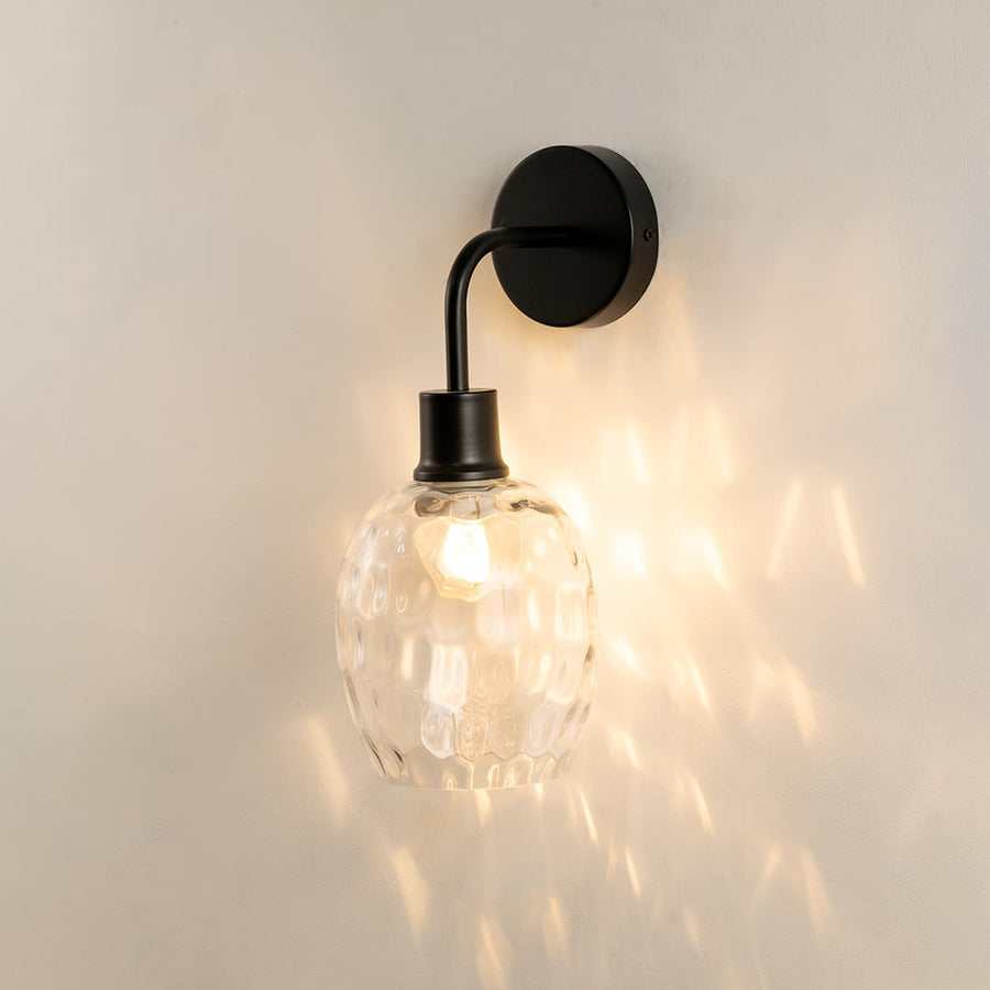 A black metal and clear glass wall light glowing warmly against a smooth pale wall spreading patterned reflections and adding stylish contemporary interior décor.