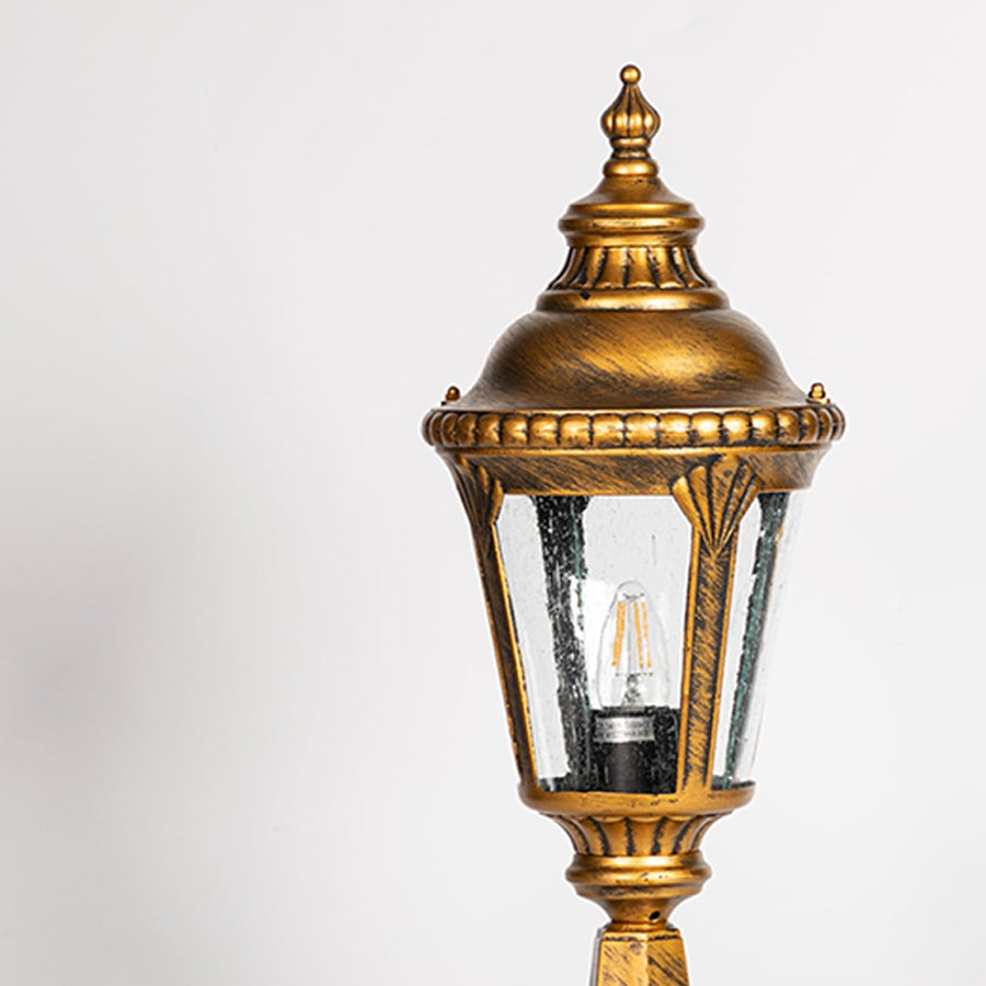 Closeup of golden pillar lamp with clear glass panels and bulb showing elegant details for outdoor gate and garden lighting.
