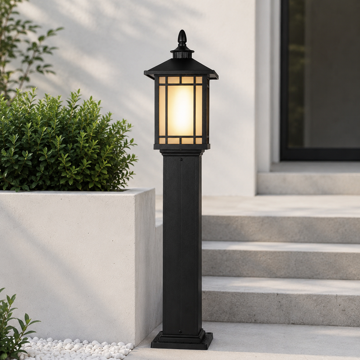 A black outdoor post lantern light with a warm glowing yellow glass shade, positioned on concrete steps next to a white planter box with green shrubs.