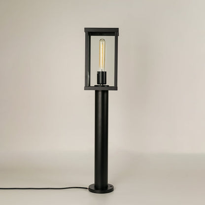 A black outdoor bollard light on a white background with the light off highlighting its clear glass panels metal frame and sturdy square base for a clean product view.