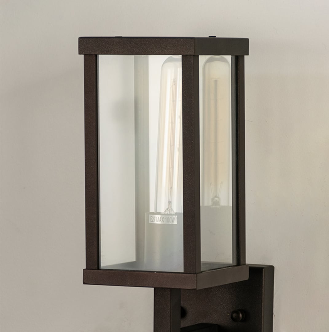Closeup view of black outdoor metal wall light showing tall clear glass panels and warm glowing bulb for modern exterior lighting.