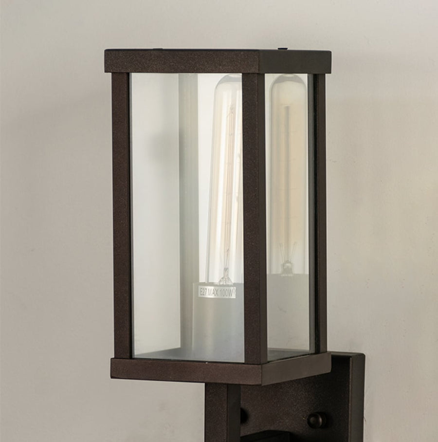 Closeup view of black outdoor metal wall light showing tall clear glass panels and warm glowing bulb for modern exterior lighting.