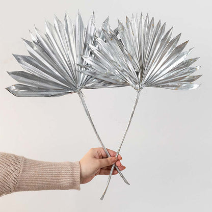 Hand holding two metallic silver painted dried palm sun fan leaves for festive luxury tabletop vase decor on white backdrop.