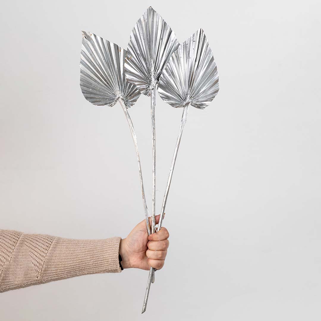 Hand holding three metallic silver painted dried palm spear leaves for festive holiday tabletop vase decor on white backdrop.