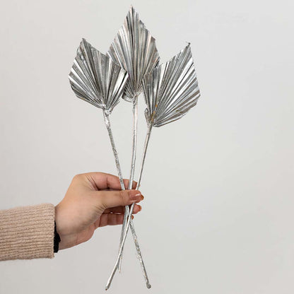 Hand holding three metallic silver painted dried palm spear leaves for festive holiday tabletop vase decor on white background.