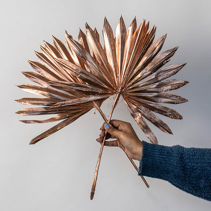 Hand holding two rose gold metallic painted dried palm sun fan leaves for festive luxury tabletop vase on white backdrop.