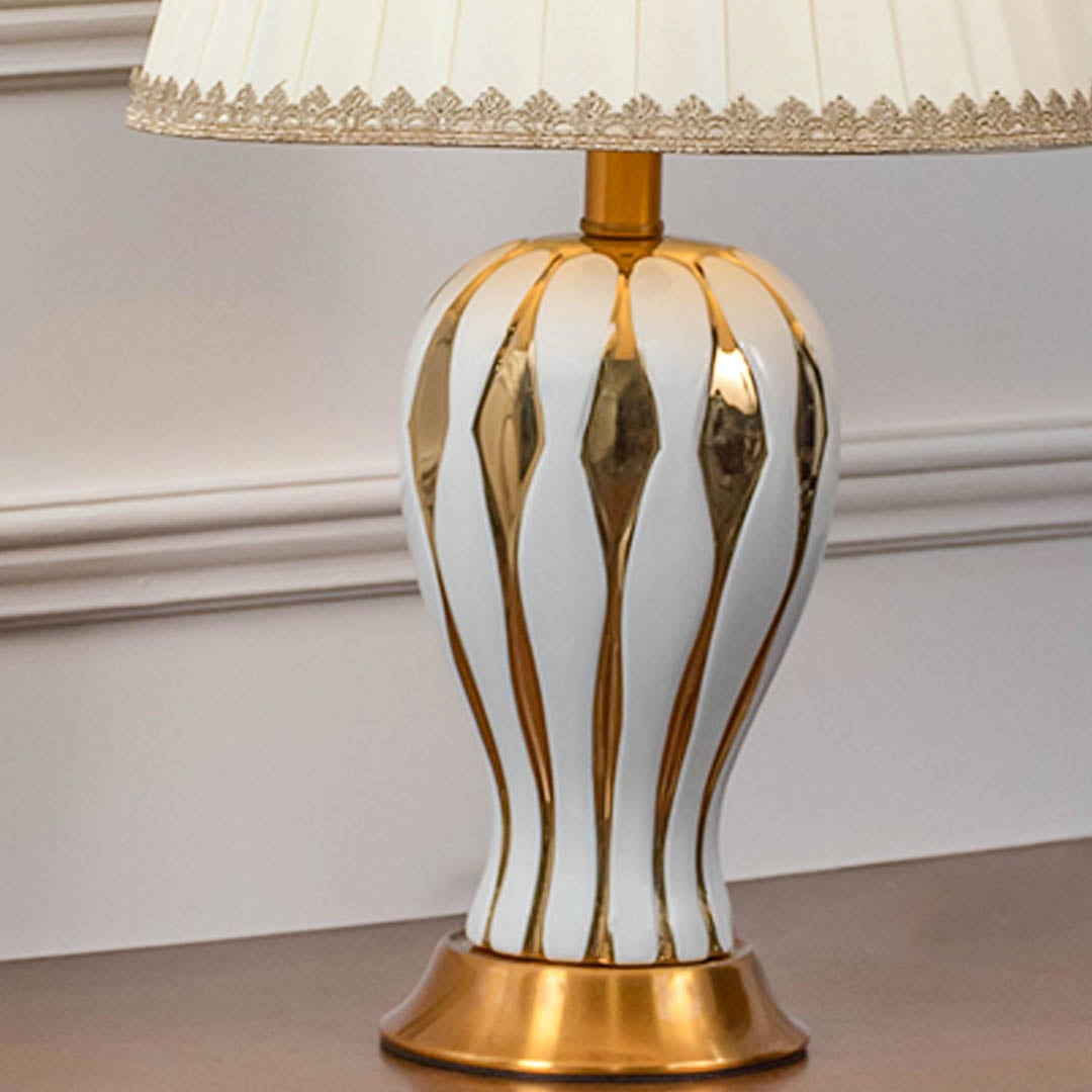 Closeup of white ceramic table lamp with golden striped ceramic base brass bottom and pleated fabric shade.