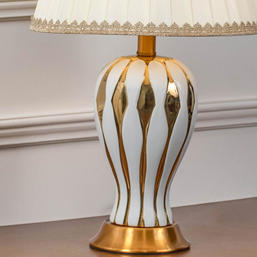 Closeup of white ceramic table lamp with golden striped ceramic base brass bottom and pleated fabric shade.