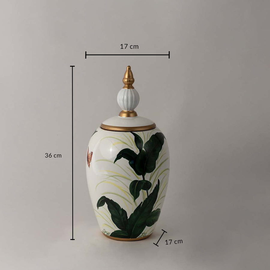 White ceramic decor jar with tropical green leaf artwork and golden lid shown with 36 cm height and 17 cm width.