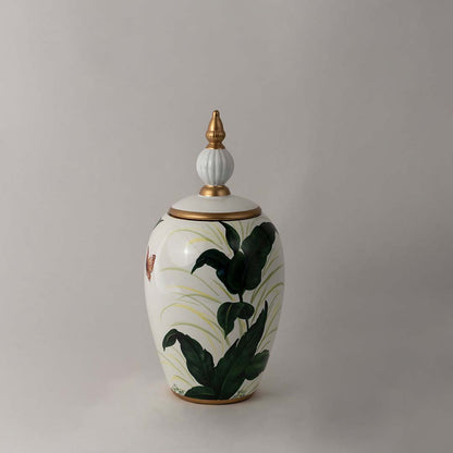 Decorative ceramic jar with green botanical leaf artwork and gold lid placed on white surface against neutral background showcasing elegant modern home decor styling.