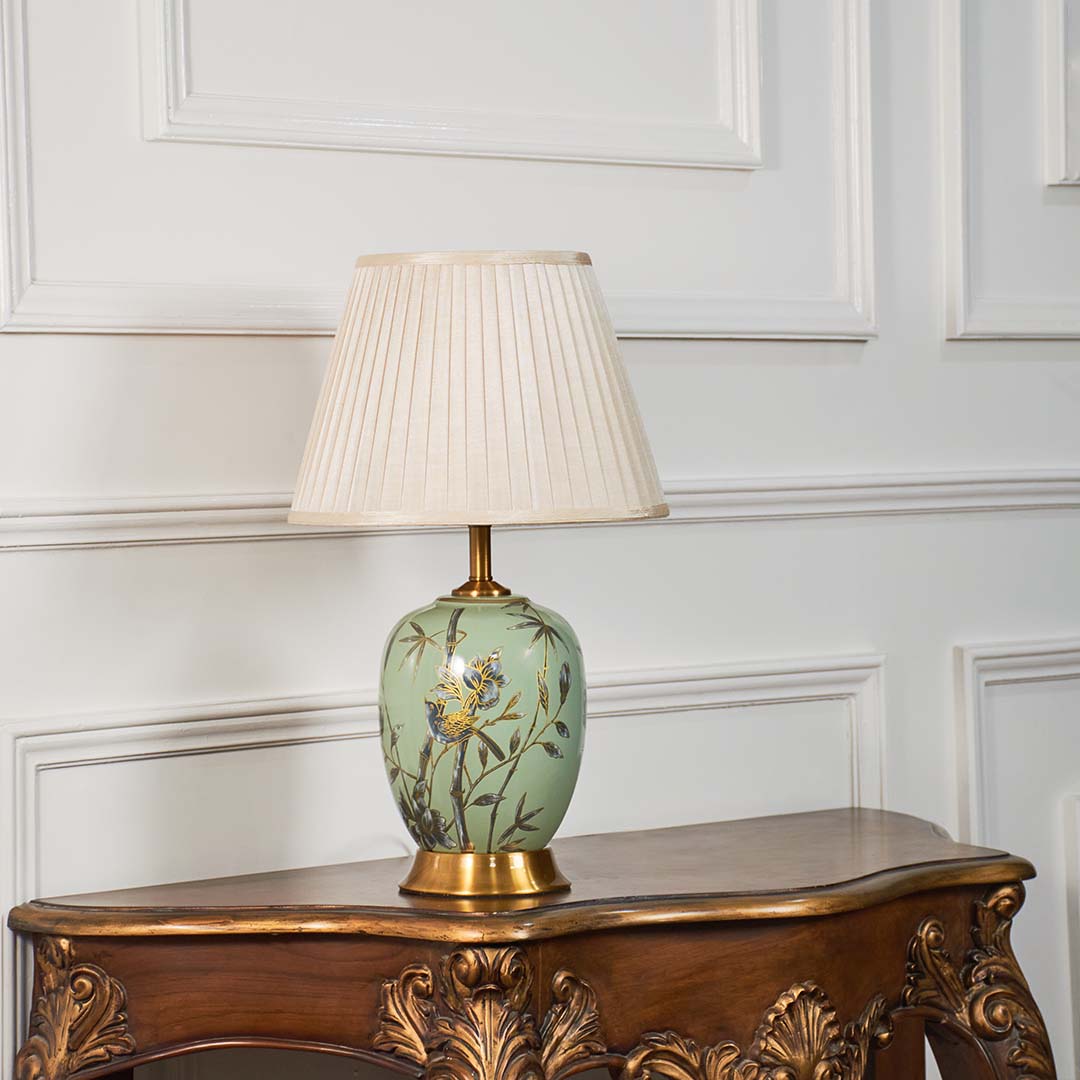 Hand-painted green floral table lamp with beige pleated lampshade glowing warmly on a clean white background.