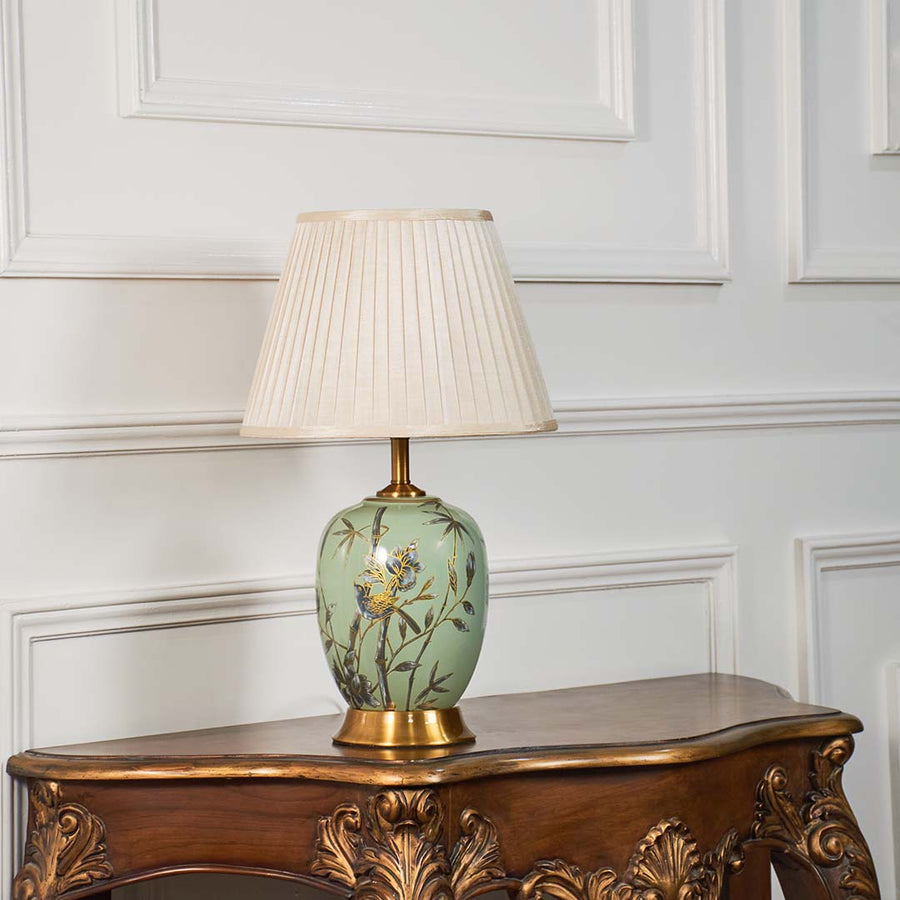 Hand-painted green floral table lamp with beige pleated lampshade glowing warmly on a clean white background.