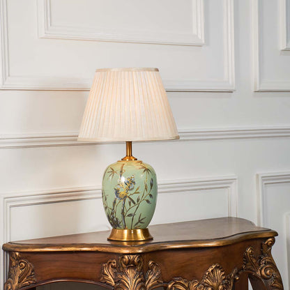 Hand-painted green floral table lamp with beige pleated lampshade glowing warmly on a clean white background.