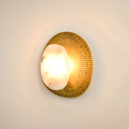 Contemporary alabaster wall sconce illuminated on white background with textured brass plate offering a soft  warm glow and sophisticated appeal.