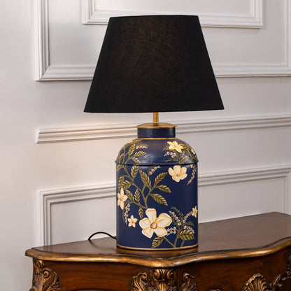 Table lamp with black fabric shade and navy ceramic base highlighted with elegant floral artwork and gold accents positioned on a wooden table against a white background.