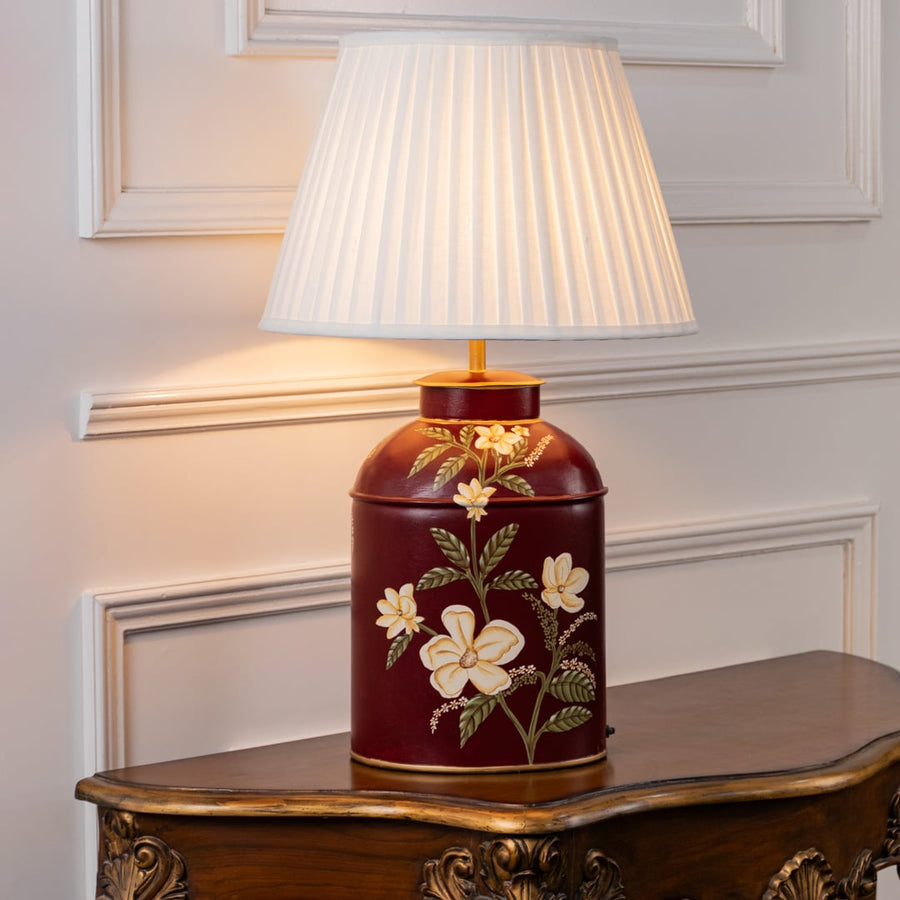 Table lamp with a white pleated shade and deep red ceramic base featuring elegant floral artwork displayed on a classic wooden table.