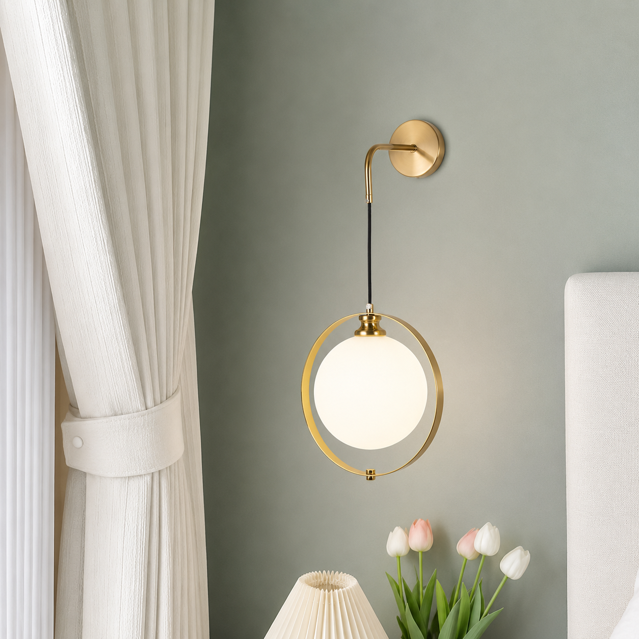 A modern wall-mounted light fixture with a circular gold-toned frame and a glowing white globe shade, installed on a muted gray wall in a styled bedroom setting with a curtain, flowers, and a lamp visible.