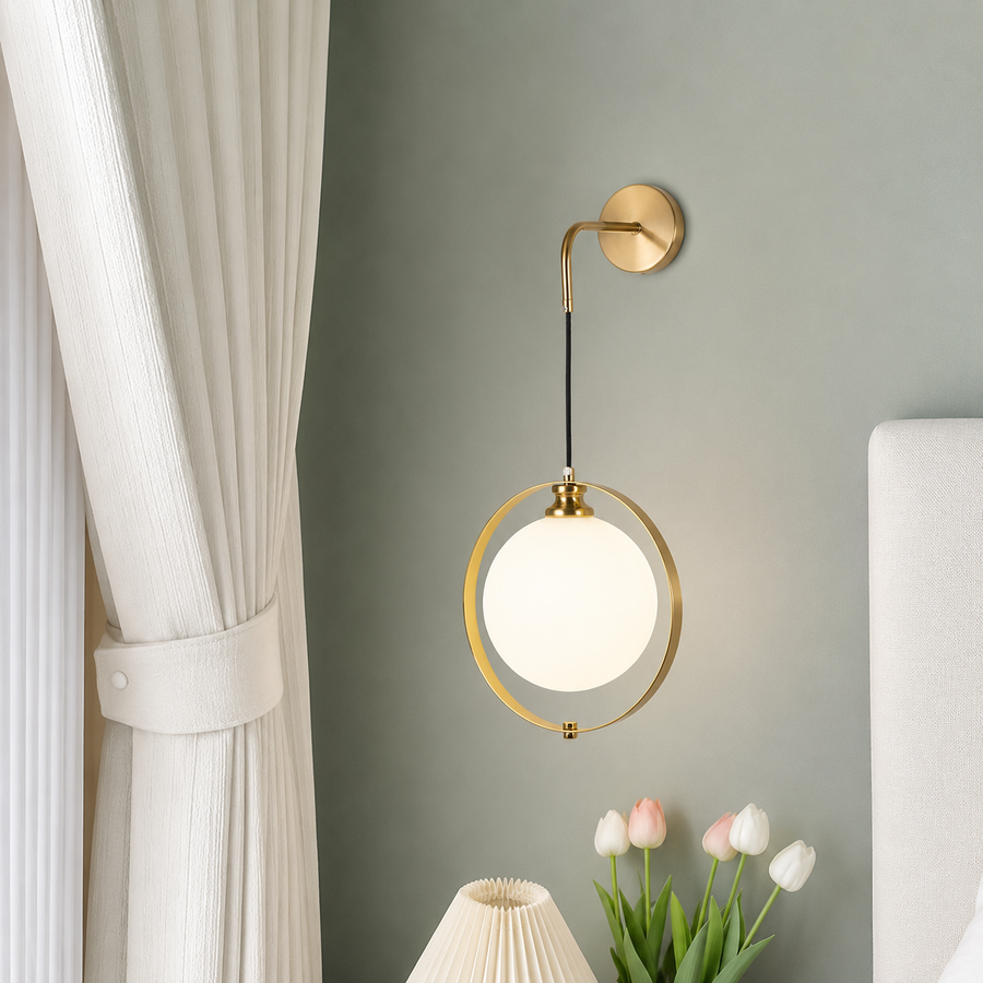 A modern wall-mounted light fixture with a circular gold-toned frame and a glowing white globe shade, installed on a muted gray wall in a styled bedroom setting with a curtain, flowers, and a lamp visible.