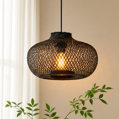 A woven rattan pendant light with a glowing warm bulb inside, hanging against a beige wall with soft window light and green plant leaves in the foreground.