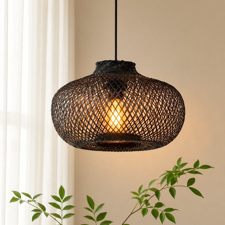 A woven rattan pendant light with a glowing warm bulb inside, hanging against a beige wall with soft window light and green plant leaves in the foreground.