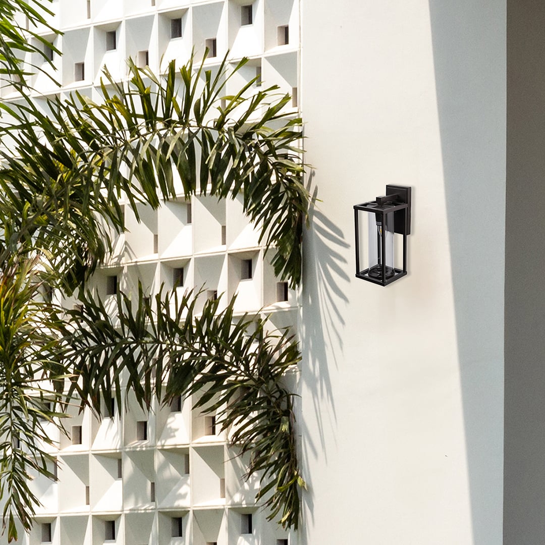 Modern outdoor wall light with black frame and clear glass shade mounted on white exterior wall near greenery.