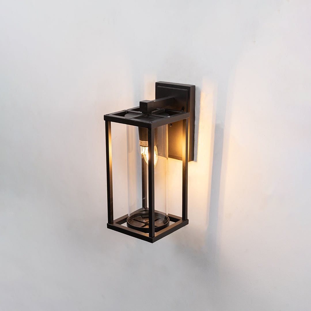 Black metal exterior wall lantern with clear glass panel housing one glowing vintage edison bulb on white background.