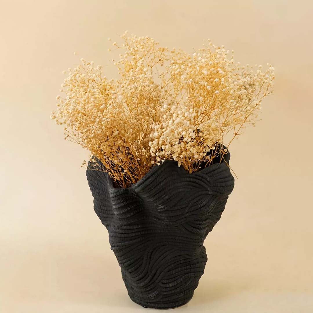Fluffy cream yellow dried flower stems bundle arranged inside an elegant modern textured black ceramic vase on living table.