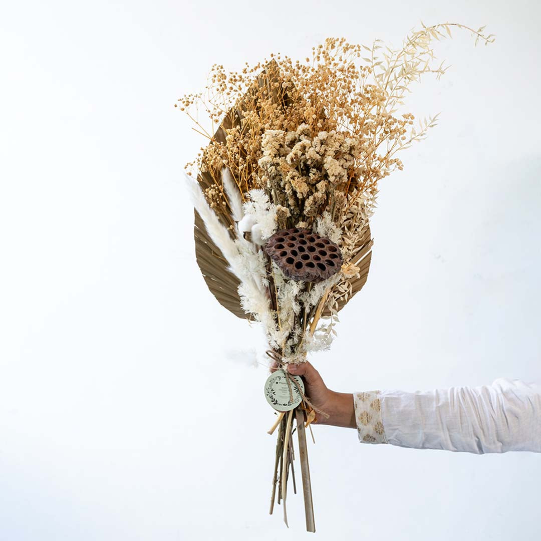 Hand holding an elegant natural beige dried floral bouquet with palm spear and grass for boho room decor on white backdrop.