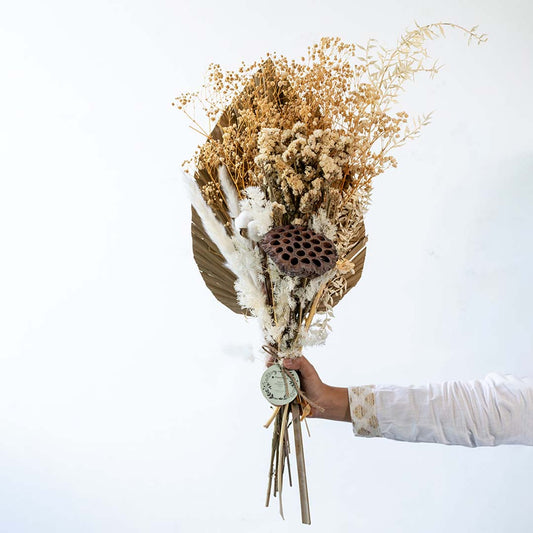 Hand holding an elegant natural beige dried floral bouquet with palm spear and grass for boho room decor on white backdrop.