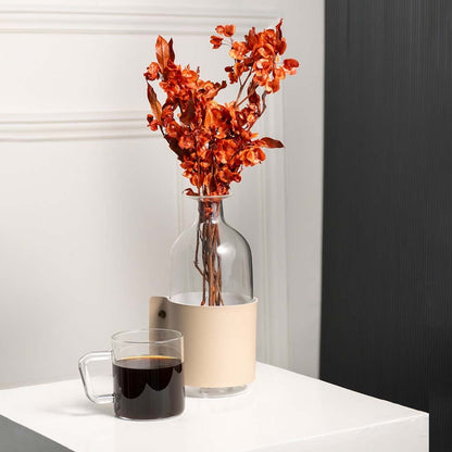 Natural dried orange botanical stems styled in a clear glass vase with beige holder beside a warm coffee mug creating a cozy modern home decor setting.