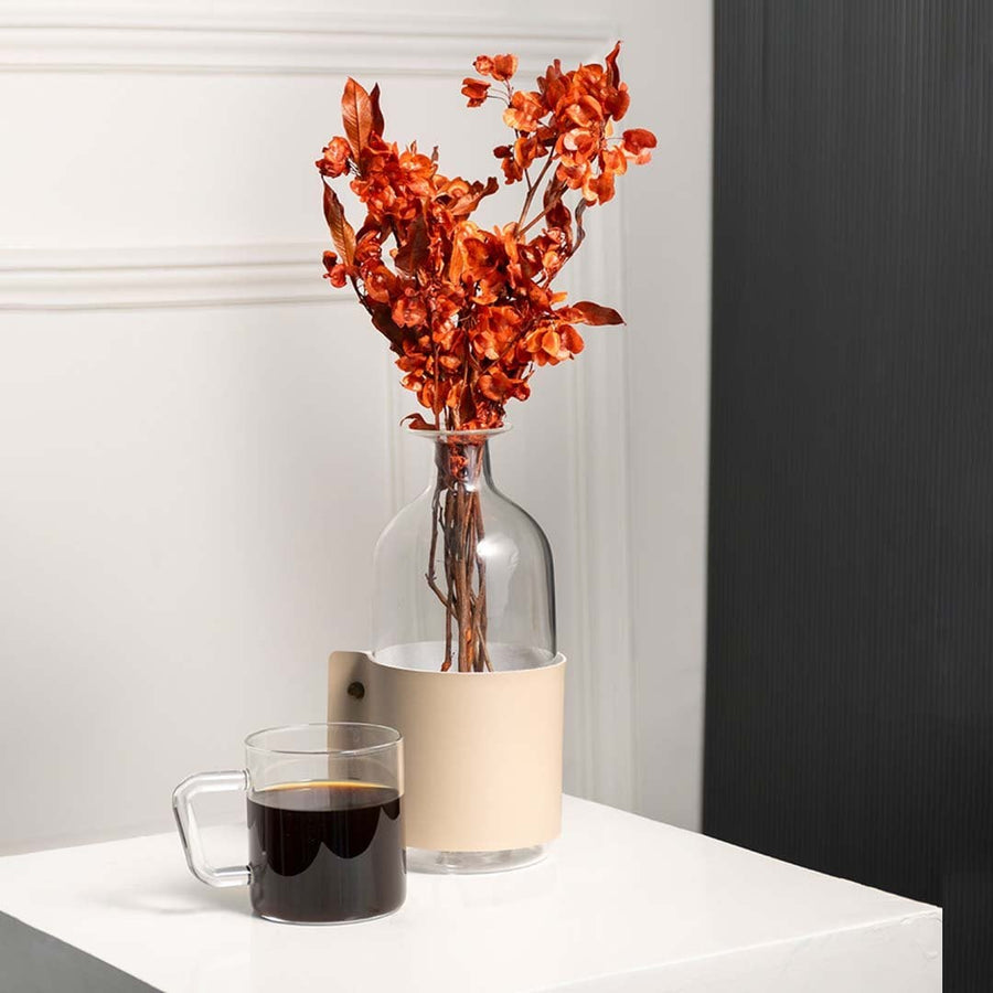 Natural dried orange botanical stems styled in a clear glass vase with beige holder beside a warm coffee mug creating a cozy modern home decor setting.