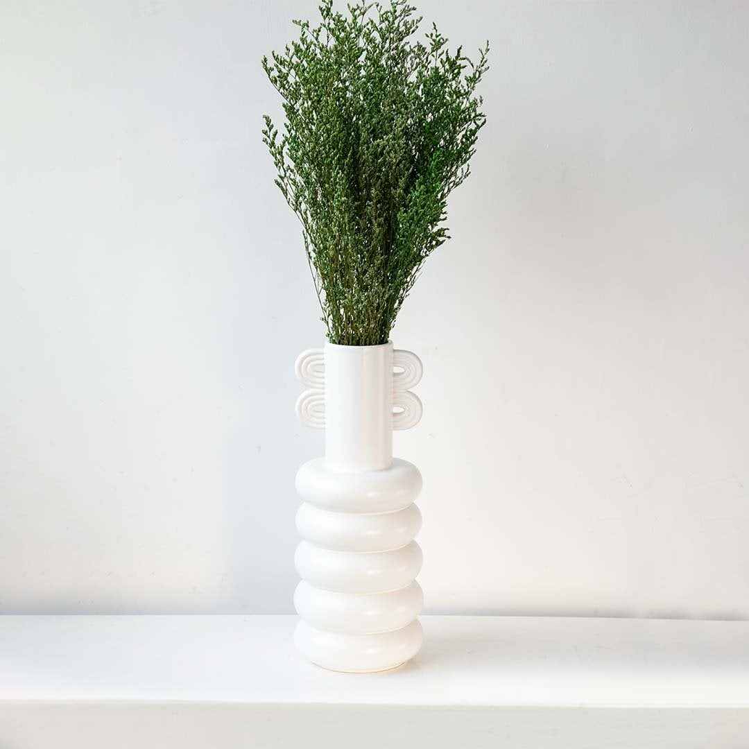 Green dried filler foliage styled in tall modern white ceramic vase against wall creating elegant lifestyle home decor.