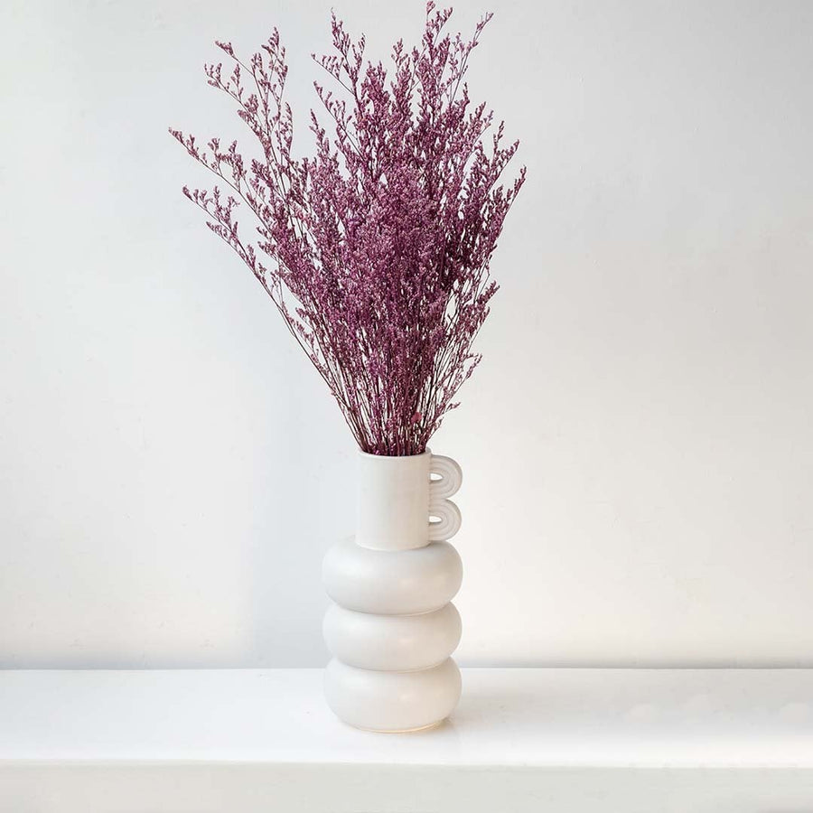 Purple dried filler foliage styled in tall sculptural white ceramic vase against wall creating modern lifestyle decor.