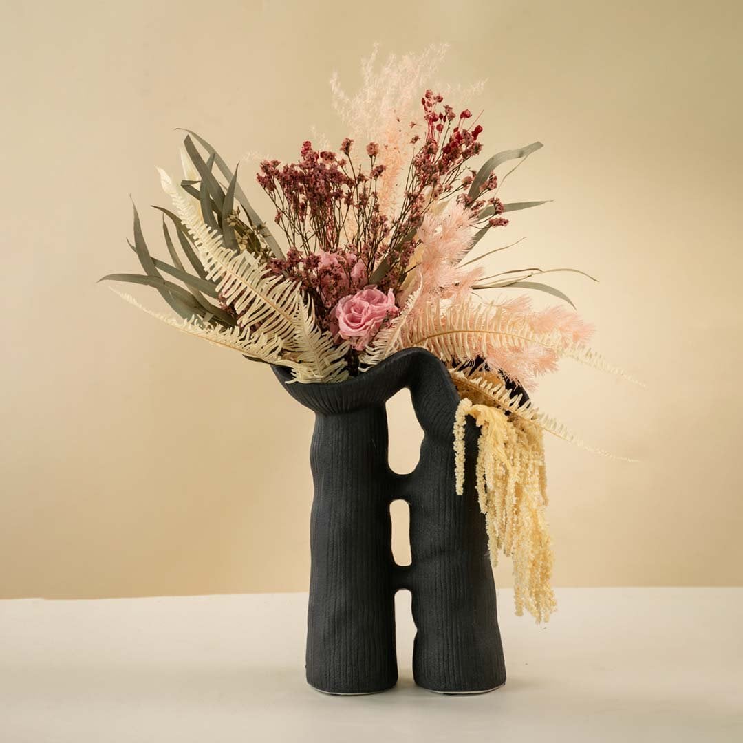 Black modern sculptural ceramic vase holding pink dried flowers and foliage arranged on a neutral white background.