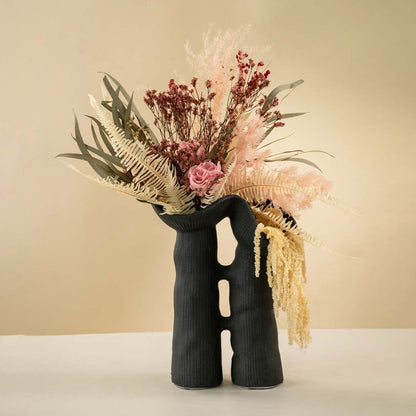 Black modern sculptural ceramic vase holding pink dried flowers and foliage arranged on a neutral white background.
