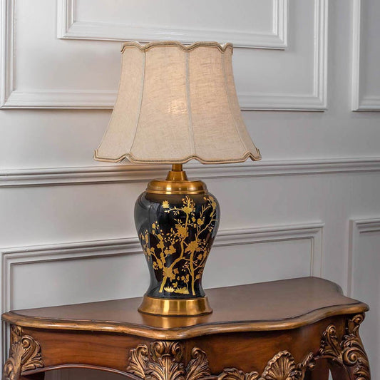 Black and gold ceramic lamp with flared beige shade turned on against white background casting soft glow perfect for classic bedroom or living room decor.