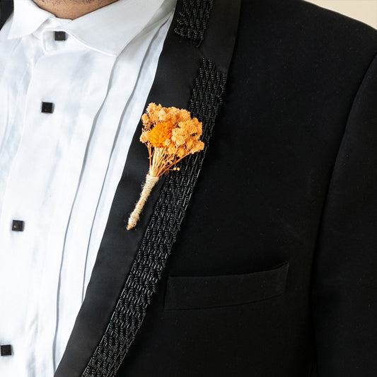 Detailed close up view of an artisanal orange fabric flower boutonniere pinned to a luxury black suit jacket lapel panel.