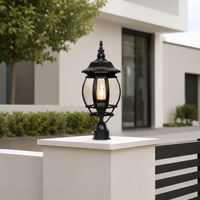 A black outdoor bollard light fixture with a warm glowing glass globe, mounted on a white concrete pillar in a modern garden setting.